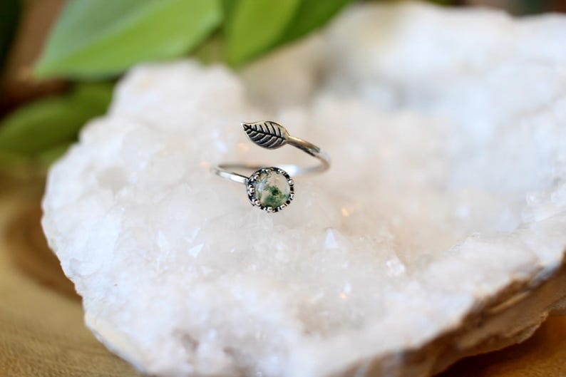 May include: A silver-toned ring with a leaf design and a round, green stone. The ring features an adjustable band and a small leaf detail. The stone has a unique, mossy appearance. The ring is set against a white, textured background.