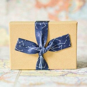 Puede incluir: Una peque&ntilde;a caja de regalo marr&oacute;n atada con una cinta azul con un patr&oacute;n de constelaci&oacute;n blanca.