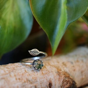 May include: A silver-toned ring with a leaf design. The ring features a small, round, green stone set in a decorative bezel. A leaf detail adorns the band, adding a nature-inspired touch. The ring is displayed on a wooden surface.