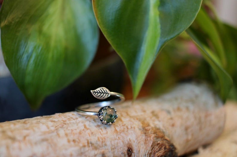 May include: A silver-toned ring with a leaf design. The ring features a small, round, green stone set in a decorative bezel. A leaf detail adorns the band, adding a nature-inspired touch. The ring is displayed on a wooden surface.