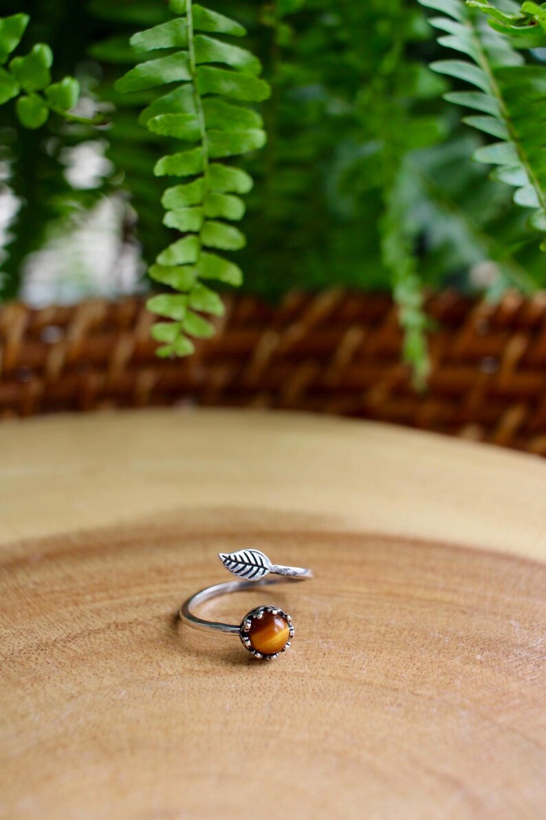 May include: A silver-toned ring with a leaf design and a round, amber-colored stone. The ring is set on a wooden surface, with green foliage in the background. The ring's design features a leaf detail and a spiral band.