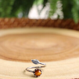 May include: A silver-toned ring with a leaf design and a round, orange-brown gemstone. The ring is set on a wooden surface, with a blurred green background.