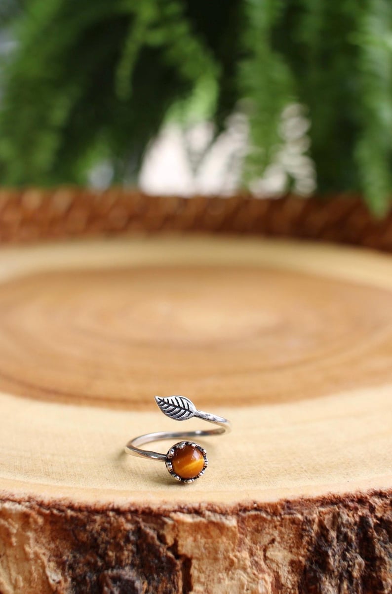 May include: A silver-toned ring with a leaf design and a round, orange-brown gemstone. The ring is set on a wooden surface, with a blurred green background.