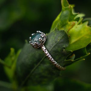 Puede incluir: Un anillo de plata con una piedra de &aacute;gata musgosa verde oscuro redonda, engastada en una montura de garras en forma de corona. La banda del anillo tiene un acabado martillado y texturizado que le da un aspecto r&uacute;stico con inclusiones naturales.