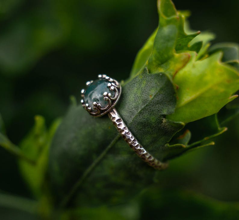 Puede incluir: Un anillo de plata con una piedra de &aacute;gata musgosa verde oscuro redonda, engastada en una montura de garras en forma de corona. La banda del anillo tiene un acabado martillado y texturizado que le da un aspecto r&uacute;stico con inclusiones naturales.