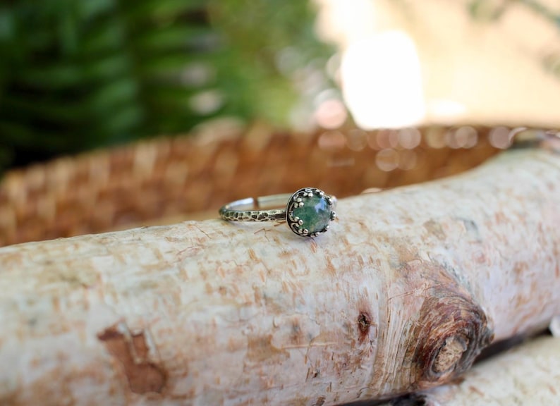 Puede incluir: Un anillo de plata con una piedra preciosa redonda de color verde moteado, engastada en un bisel de metal texturizado con cuentas. La banda del anillo tiene un acabado martillado e irregular, lo que le da un aspecto t&aacute;ctil a la estructura.