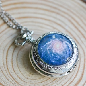 Puede incluir: Un reloj de bolsillo plateado con una cadena. La esfera del reloj tiene una imagen azul y blanca de una galaxia.