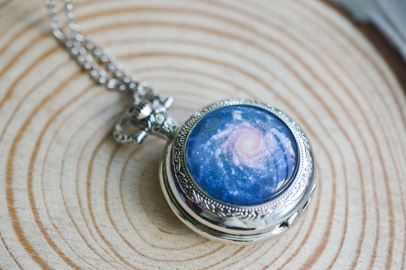 Puede incluir: Un reloj de bolsillo plateado con una cadena. La esfera del reloj tiene una imagen azul y blanca de una galaxia.