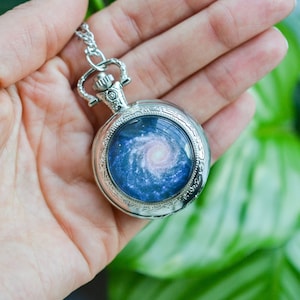 Puede incluir: Un reloj de bolsillo plateado con una cadena, que presenta un dise&ntilde;o de galaxia con una nebulosa azul y rosa giratoria.