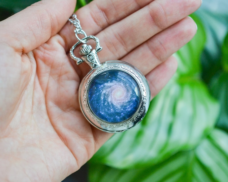 Puede incluir: Un reloj de bolsillo plateado con una cadena, que presenta un dise&ntilde;o de galaxia con una nebulosa azul y rosa giratoria.