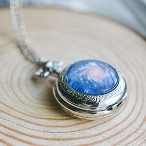Puede incluir: Un reloj de bolsillo plateado con un dise&ntilde;o de galaxia. La esfera del reloj presenta una galaxia azul y rosa con una nebulosa giratoria.