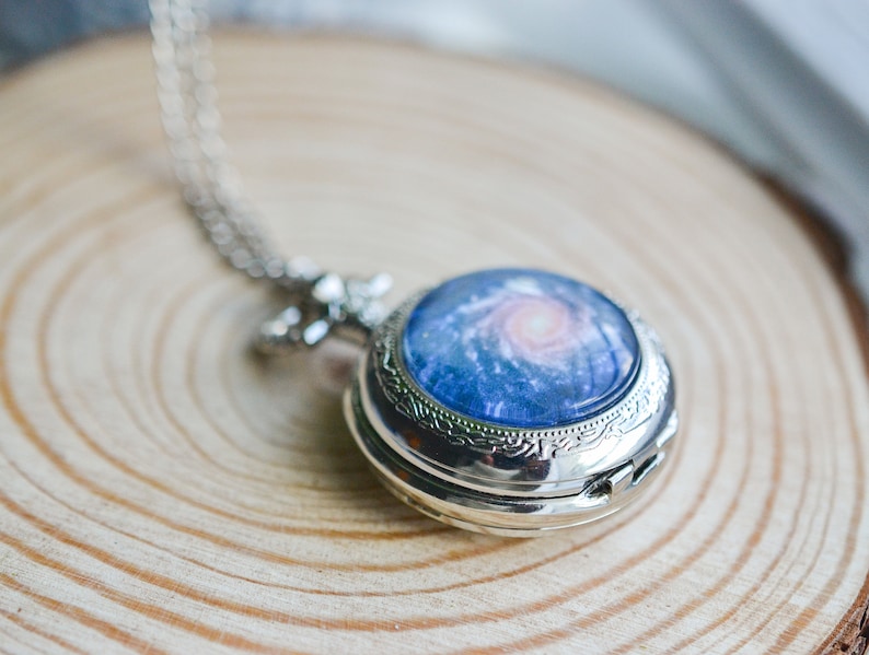 Puede incluir: Un reloj de bolsillo plateado con un dise&ntilde;o de galaxia. La esfera del reloj presenta una galaxia azul y rosa con una nebulosa giratoria.