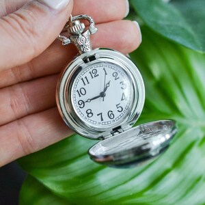 Puede incluir: Un reloj de bolsillo plateado con una esfera blanca y agujas negras. El reloj est&aacute; abierto y el interior es visible.