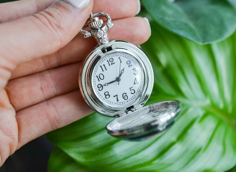 Puede incluir: Un reloj de bolsillo plateado con una esfera blanca y agujas negras. El reloj est&aacute; abierto y el interior es visible.