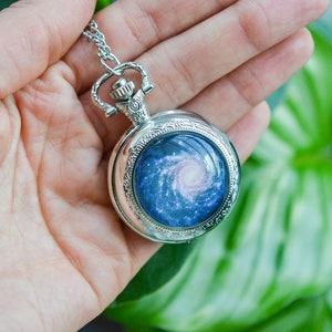Puede incluir: Un reloj de bolsillo plateado con una cadena, que presenta un dise&ntilde;o de galaxia en la esfera. La galaxia es una nebulosa azul y rosa que gira.