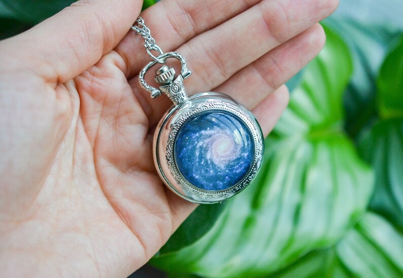 Puede incluir: Un reloj de bolsillo plateado con una cadena, que presenta un dise&ntilde;o de galaxia en la esfera. La galaxia es una nebulosa azul y rosa que gira.