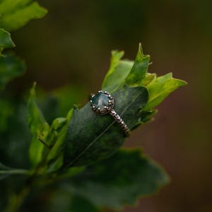 Puede incluir: Un anillo de plata con una piedra preciosa redonda, transl&uacute;cida y de color verde musgo, engastada en una montura de garras en forma de corona. La banda del anillo tiene un acabado martillado y texturizado, con un aspecto t&aacute;ctil.