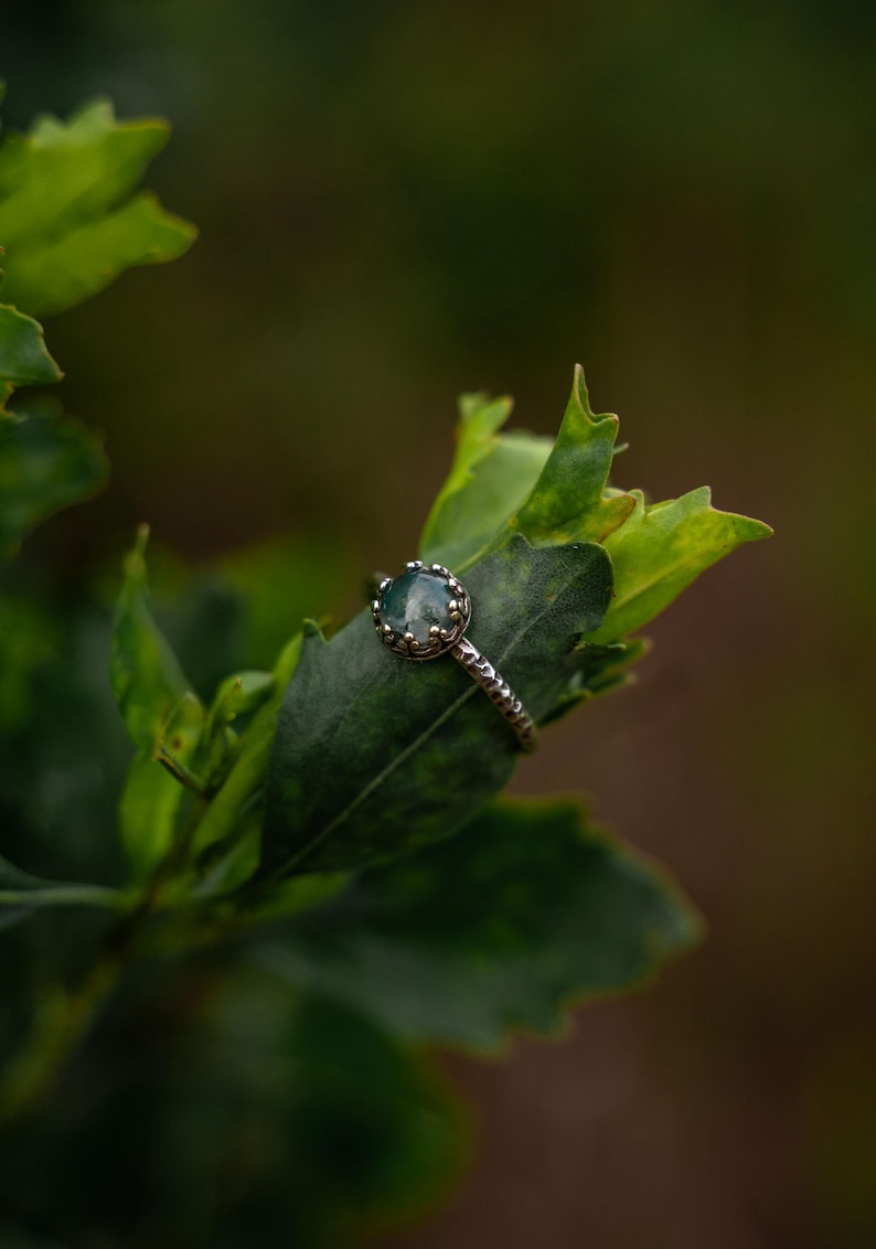 Puede incluir: Un anillo de plata con una piedra preciosa redonda, transl&uacute;cida y de color verde musgo, engastada en una montura de garras en forma de corona. La banda del anillo tiene un acabado martillado y texturizado, con un aspecto t&aacute;ctil.