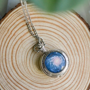 Puede incluir: Un collar con un reloj de bolsillo plateado con un dise&ntilde;o de galaxia azul y blanco en la esfera.