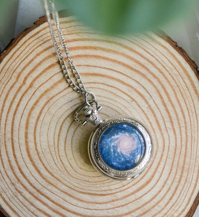 Puede incluir: Un collar con un reloj de bolsillo plateado con un dise&ntilde;o de galaxia azul y blanco en la esfera.