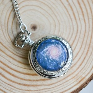 Puede incluir: Un reloj de bolsillo plateado con una cadena. La esfera del reloj presenta un dise&ntilde;o de galaxia azul y rosa.