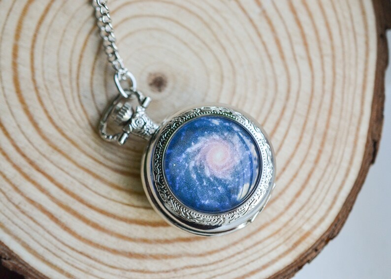 Puede incluir: Un reloj de bolsillo plateado con una cadena. La esfera del reloj presenta un dise&ntilde;o de galaxia azul y rosa.