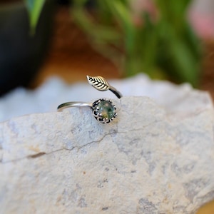 May include: A silver-toned ring with a leaf and gemstone design. The ring features a small, round, green gemstone set in a decorative bezel. A delicate leaf detail extends from the ring band, adding a natural element. The ring is displayed on a light-colored stone.