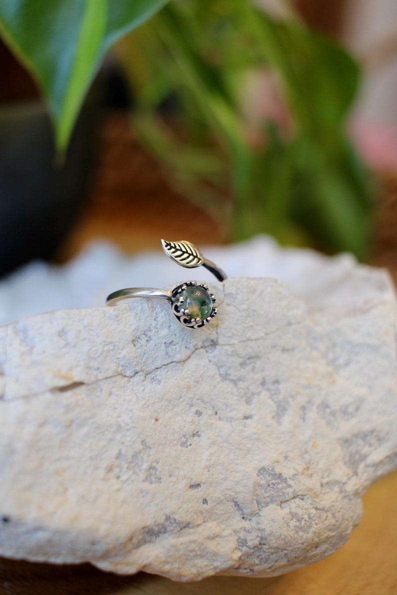 May include: A silver-toned ring with a leaf and gemstone design. The ring features a small, round, green gemstone set in a decorative bezel. A delicate leaf detail extends from the ring band, adding a natural element. The ring is displayed on a light-colored stone.