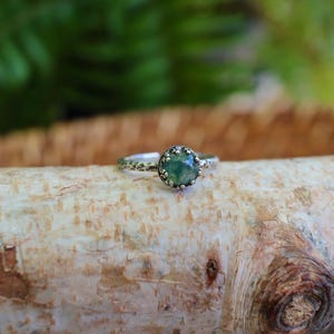 Puede incluir: Un anillo de plata con una piedra preciosa redonda de color verde musgo engastada en una montura de garras en forma de corona. La banda del anillo tiene un acabado martillado y texturizado, que le da un aspecto rugoso que contrasta con la piedra.