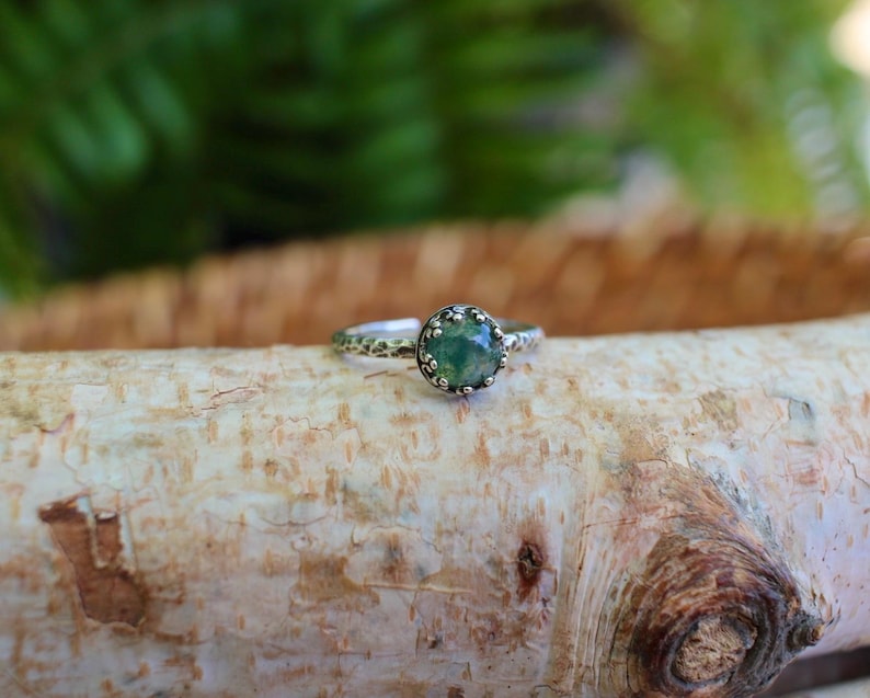 Puede incluir: Un anillo de plata con una piedra preciosa redonda de color verde musgo engastada en una montura de garras en forma de corona. La banda del anillo tiene un acabado martillado y texturizado, que le da un aspecto rugoso que contrasta con la piedra.