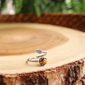 May include: A silver-toned ring with a leaf design and a round, amber-colored stone. The ring is set on a wooden surface with a natural edge, showcasing the ring's organic theme. The ring's design is reminiscent of nature.