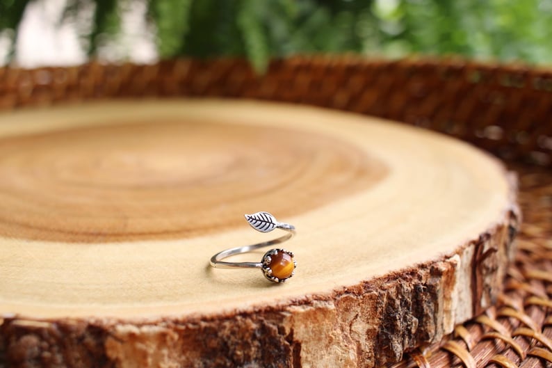 May include: A silver-toned ring with a leaf design and a round, amber-colored stone. The ring is set on a wooden surface with a natural edge, showcasing the ring's organic theme. The ring's design is reminiscent of nature.