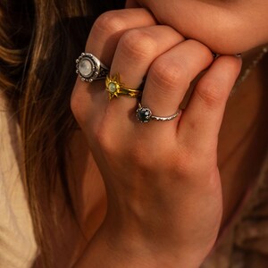 Puede incluir: Tres anillos en los dedos. Un anillo de plata presenta una piedra redonda iridiscente con borde de cuentas. Un anillo de oro muestra un dise&ntilde;o de estrella con una piedra central. Un anillo de plata fino y texturizado tiene una piedra oscura.