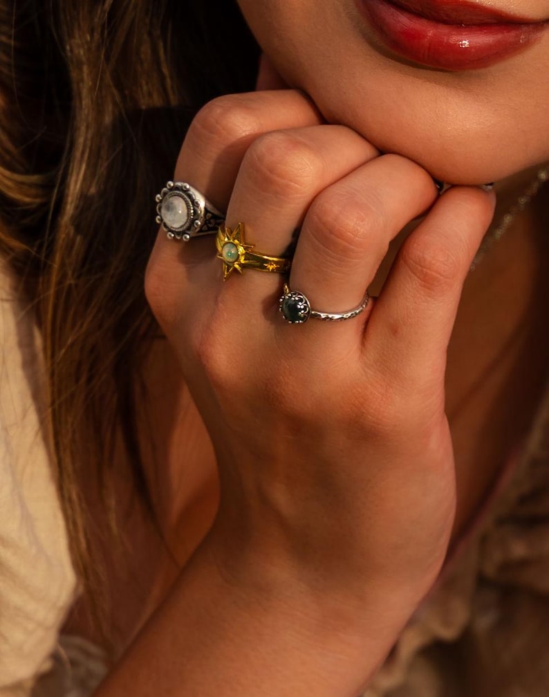 Puede incluir: Tres anillos en los dedos. Un anillo de plata presenta una piedra redonda iridiscente con borde de cuentas. Un anillo de oro muestra un dise&ntilde;o de estrella con una piedra central. Un anillo de plata fino y texturizado tiene una piedra oscura.