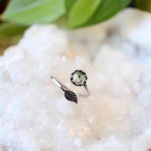 May include: A silver-toned ring with a leaf accent and a round, green stone. The ring's design features an open band with a detailed leaf on one side and a bezel-set stone on the other. The stone has a mossy appearance.