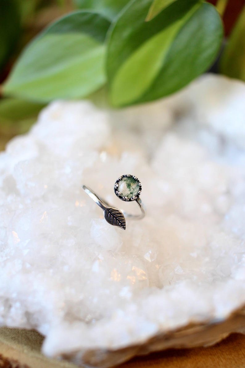 May include: A silver-toned ring with a leaf accent and a round, green stone. The ring's design features an open band with a detailed leaf on one side and a bezel-set stone on the other. The stone has a mossy appearance.