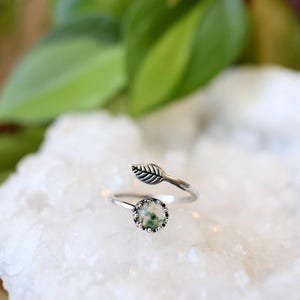 May include: A silver ring with a leaf design and a round, moss agate stone. The ring features a delicate leaf detail on the band and a bezel-set stone with green and white inclusions. The ring is set on a white surface.