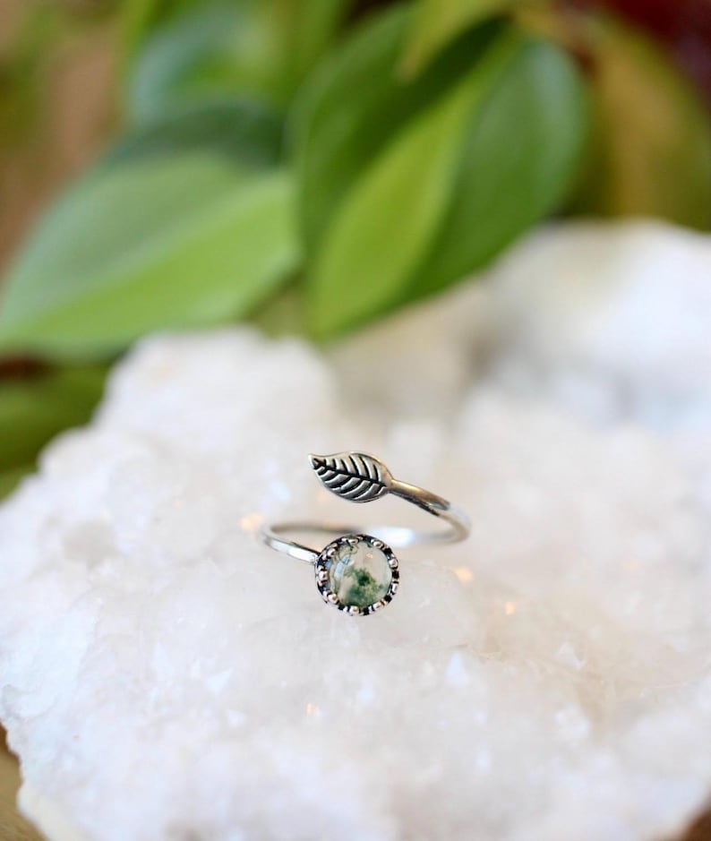 May include: A silver ring with a leaf design and a round, moss agate stone. The ring features a delicate leaf detail on the band and a bezel-set stone with green and white inclusions. The ring is set on a white surface.
