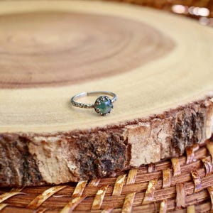 Puede incluir: Un anillo de tono plateado con una peque&ntilde;a piedra preciosa redonda de color verde moteado, engastada en un bisel texturizado en forma de corona. La banda del anillo presenta una textura martelada y con hoyuelos en toda su circunferencia.