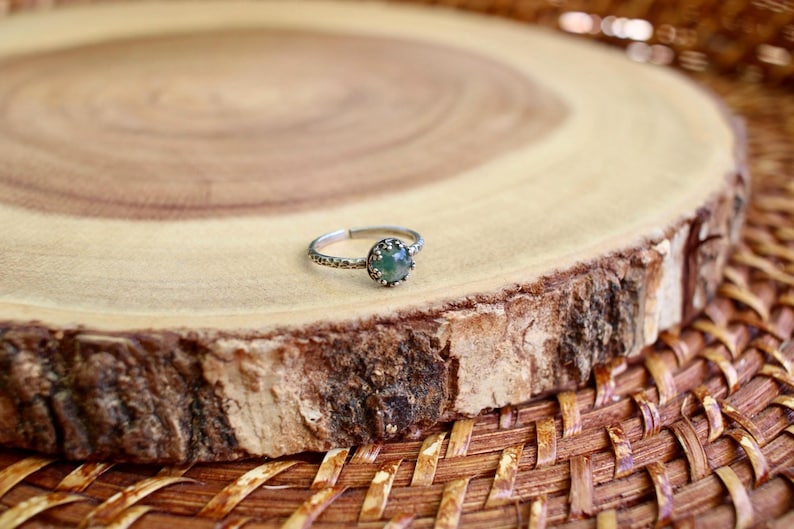 Puede incluir: Un anillo de tono plateado con una peque&ntilde;a piedra preciosa redonda de color verde moteado, engastada en un bisel texturizado en forma de corona. La banda del anillo presenta una textura martelada y con hoyuelos en toda su circunferencia.