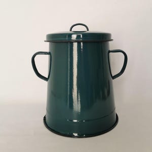 Pode incluir: Um recipiente de esmalte em tom verde-azulado escuro com corpo cilíndrico, tampa a condizer com uma pequena pega e duas pegas laterais. O recipiente apresenta um acabamento liso e brilhante com um rebordo escuro na tampa e na base.