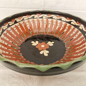May include: A round ceramic bowl with a glossy black interior and exterior. The interior features a central orange flower with cream petals, surrounded by a ring of vertical orange stripes and a decorative cream and orange scalloped border.