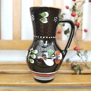 Könnte beinhalten: Ein dunkelbrauner Keramikkrug mit einem handgemalten Blumenmotiv in Weiß, Grün und Orange. Das Gefäß hat einen ausgestellten Rand, einen geschwungenen Henkel und horizontale Streifen in Weiß und Orange an der Basis sowie am Halsbereich.