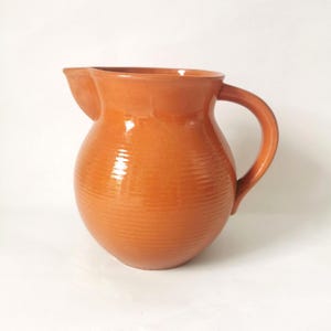 May include: A terracotta-colored ceramic pitcher with a rounded body, a wide pouring spout, and a curved handle. The surface of the pitcher features subtle horizontal ridges that wrap around the entire circumference of the vessel.