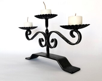 Puede incluir: Un candelabro de metal negro con tres platos festoneados para velas y velas de té blancas. La base es rectangular con extremos angulados y los brazos curvos presentan detalles en espiral que sostienen las velas.