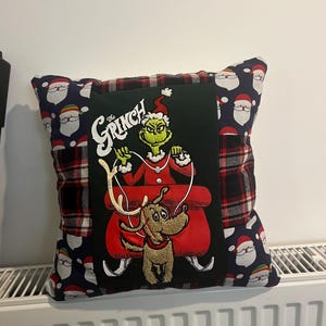 May include: A square decorative cushion featuring a central black panel with an embroidered graphic of The Grinch in a red sleigh with a dog. The side panels are made of a navy blue fabric with a repeating Santa Claus pattern and red plaid fabric.