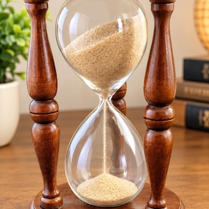 Puede incluir: Un reloj de arena con marco de madera marrón oscuro, pilares torneados y bases circulares en la parte superior e inferior. Los bulbos de vidrio transparente contienen arena beige fina, fluyendo a través del estrecho cuello central.
