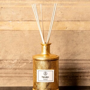 May include: A gold-coloured cylindrical reed diffuser bottle with a textured metallic finish. The bottle features a white rectangular label with the text Marceline Home Leaf Decor and Sarah. Five thin, light-coloured wooden reeds extend from the top.