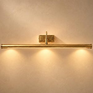 Könnte beinhalten: Eine horizontale Wandleuchte für Bilder mit gebürstetem Messing-Finish. Die lange, zylindrische Lampe verfügt über eine zentrale rechteckige Montageplatte und strahlt ein warmes, nach unten gerichtetes Licht über die gesamte Länge aus.