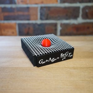 May include: A black square display piece featuring a white grid pattern on top that curves downward to hold a small red sphere. The side of the black base is printed with the white Einstein field equation Gμν + Λgμν = 8πGTμν/c4.